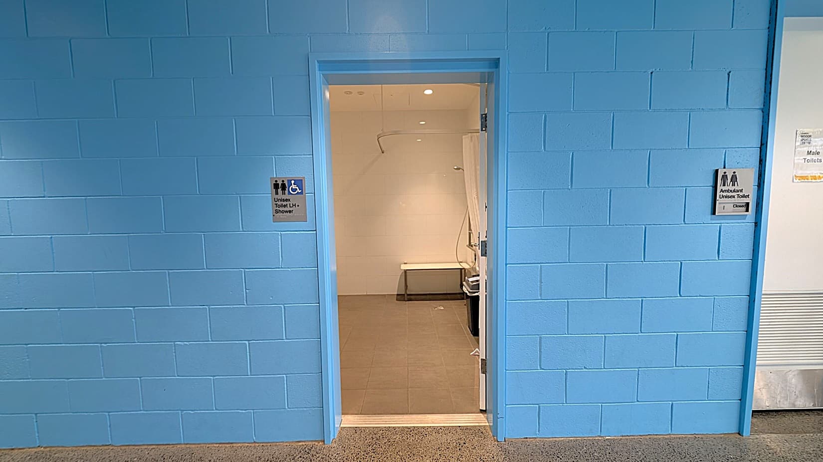 A blue brick wall with an open doorway in the centre. The doorway leads to a unisex toilet with a shower, indicated by a sign on the left side of the door. Inside, there is a bench and a showerhead visible. To the right of the doorway, there is another sign for an ambulant unisex toilet, which is marked as closed. The floor outside the doorway is speckled, and the interior floor is tiled. The ceiling inside the room has recessed lighting.