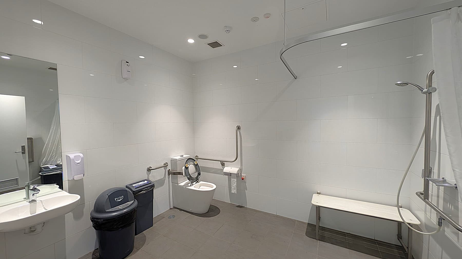 A spacious, accessible bathroom with white tiled walls and grey tiled floor. On the left, there is a sink with a mirror above it, a soap dispenser, a black bin labelled "PUSH," and a smaller black bin. In the middle, there is a toilet with grab bars on both sides and a toilet paper holder. On the right, there is a shower area with a handheld showerhead and a white bench. The ceiling has recessed lighting and a ventilation system.