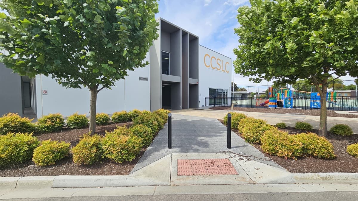 Copper Coast Sport and Leisure Centre is a large, white, and grey industrial building with a bold yellow-orange “CCSLC” sign on the left and front of the façade. The centre has glass entry doors and windows with white metal frames. Close to the left of the building are accessible car parking spaces, trees, and gardens with bushes maintained at a similar height. Right of the pavement leading to the entrance is a black, low, iron fence.