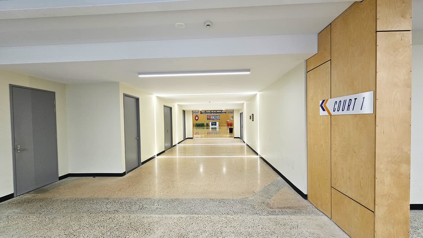 Corridor leading to Court One. A white “Court 1” sign with orange and blue accents is affixed on a wooden right wall. Four grey manual doors with stainless steel door handles are accessible on the left side. The floor transitions from light grey to light brown natural stone flooring. Long fluorescent lights reflect on the wall and floor, illuminating the corridor. A fire sprinkler can be found on the ceiling beside the white circular ventilation grille for air conditioning.