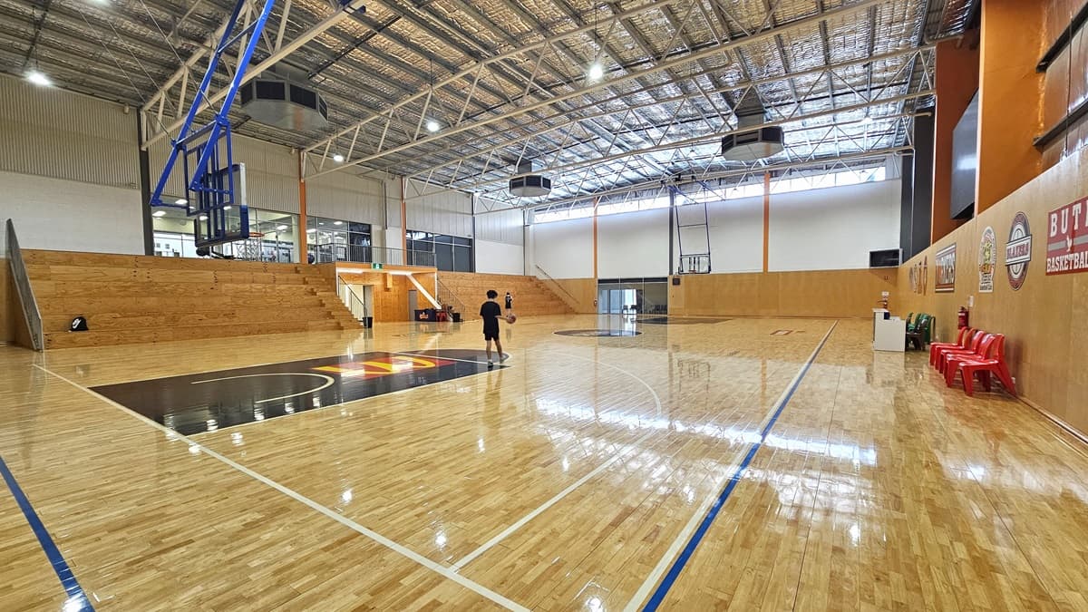A view on the edge of the expansive court. Two individuals are visible inside the court. Adjacent to the exposed steel beams and the insulated ceiling, fluorescent lights illuminate the court. The court surface features game floor markings in blue, white, and black. Between the tiered timbered seating on the left is the exit sign. On the right are the plastic chairs, posters, and a white table.