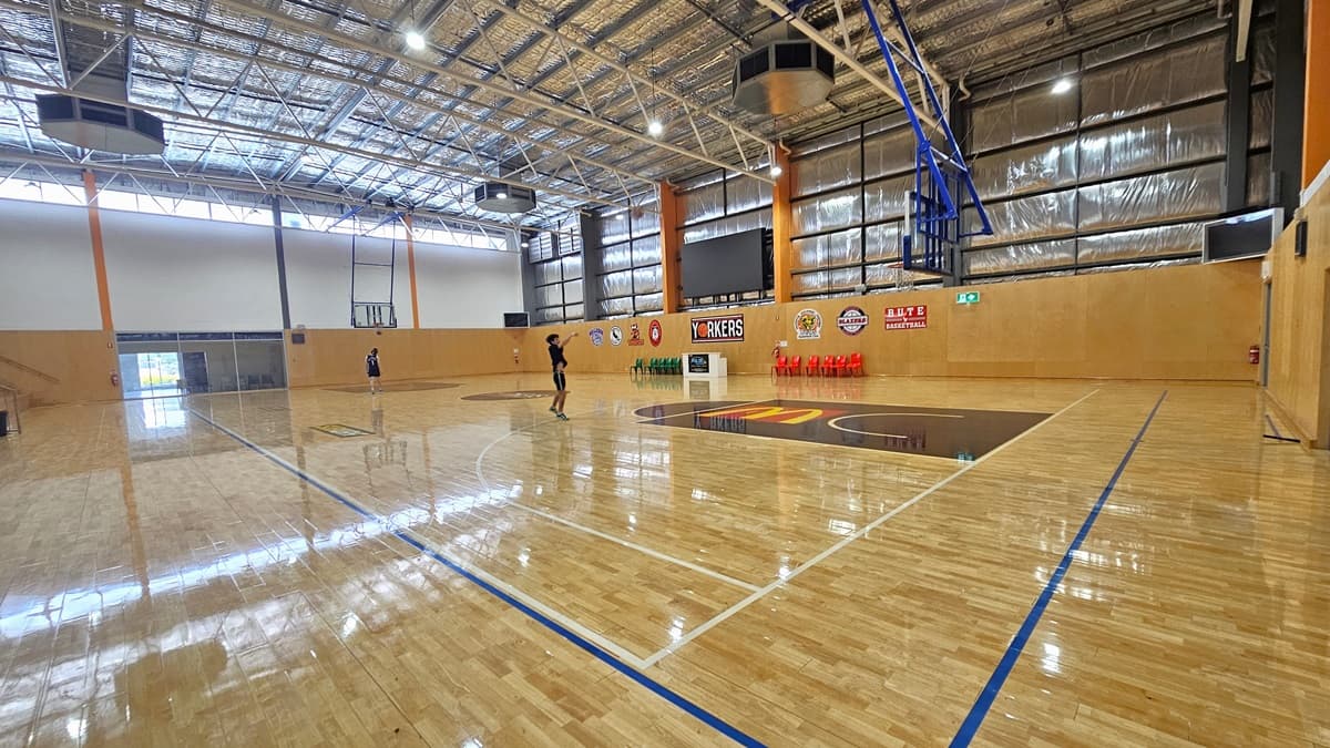 A different angle inside the basketball court. Near the exposed steel beams and the insulated foams above the ceiling are three heptagon-shaped black structures. Inside the court, two individuals are visible, with one of the individuals shooting a ball. Above the right wall filled with logos is a large digital screen mounted on the wall. Fire extinguishers are placed on the corners close to the doors.