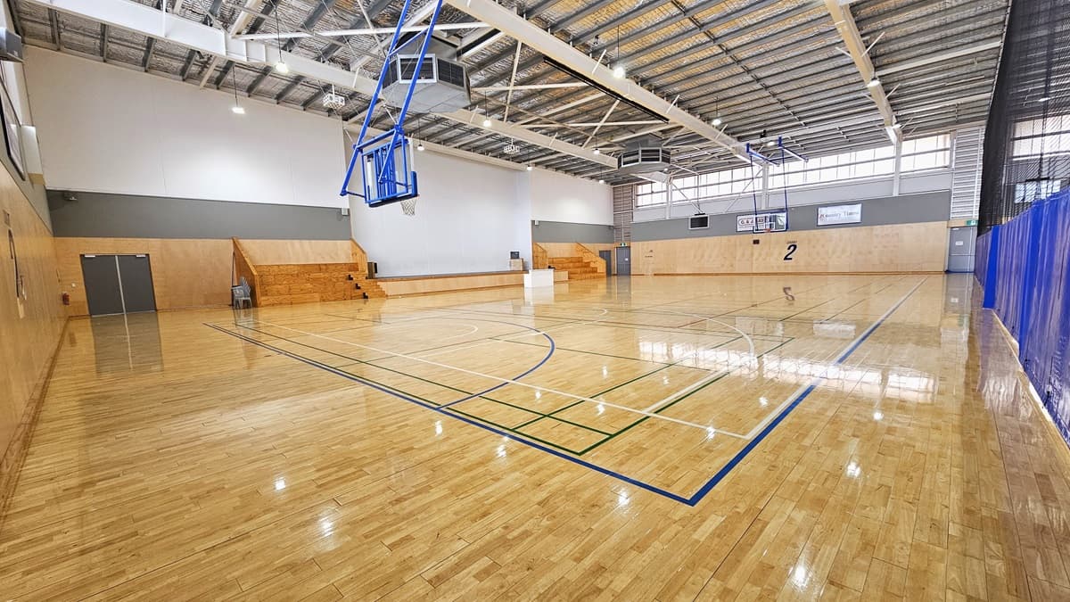 Inside the spacious Court Two. Two tiered timber seatings are visible on the left, where a sizable stage accessible through a one-stepped timbered stair is situated in the middle. On the left and right side of the court, a “2” sign is affixed on the walls. Blue, green, red, and white markings dominate the timbered floor surface.