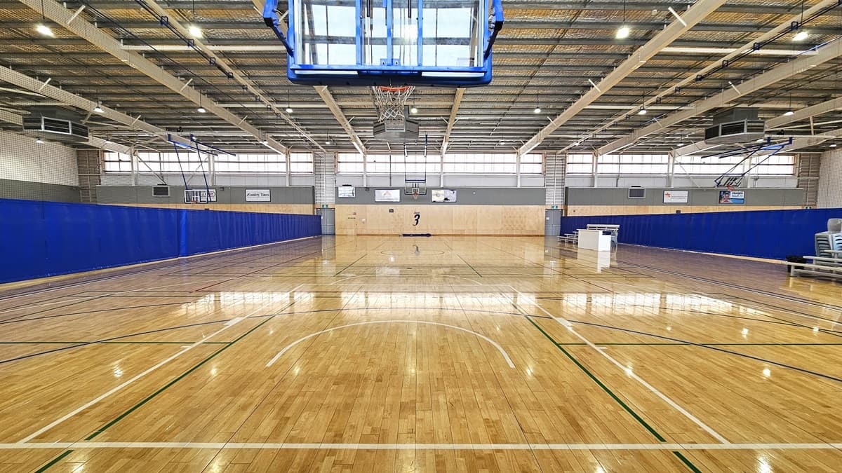 Inside the expansive Court Three. A number 3 is affixed on the court’s walls further ahead. Lengthy blue barriers are prominent on the left and right sides of the court. Two basketball hoops with supporting rails in blue facing each other are hanging on the court. On the right, stepped benches are situated, with one of the benches located next to a white table.