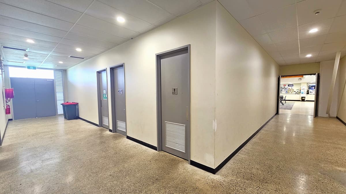 Pathway leading to the accessible bathrooms. Three accessible bathrooms equipped with gray manual doors and signs are prominent in the middle. On the left, red and black colored trash bins are placed next to the exit doors. A red fire hose is on the left side of the exit area. A pathway leading to the reception area is on the right.