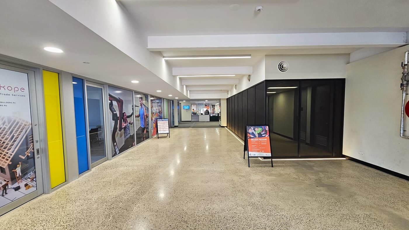 Spacious corridor with a light brown natural stone floor leading to the reception area. On both sides of the corridor are two black foldable sidewalk sign boards. On the left are the sports therapy clinician offices that have large advertisements covering their glass windows. On the right side is a glass-tinted section. Lined on the white wall above it are white circular ventilation grille for air conditioning. On the left wall of the reception desk is a small flat TV screen.