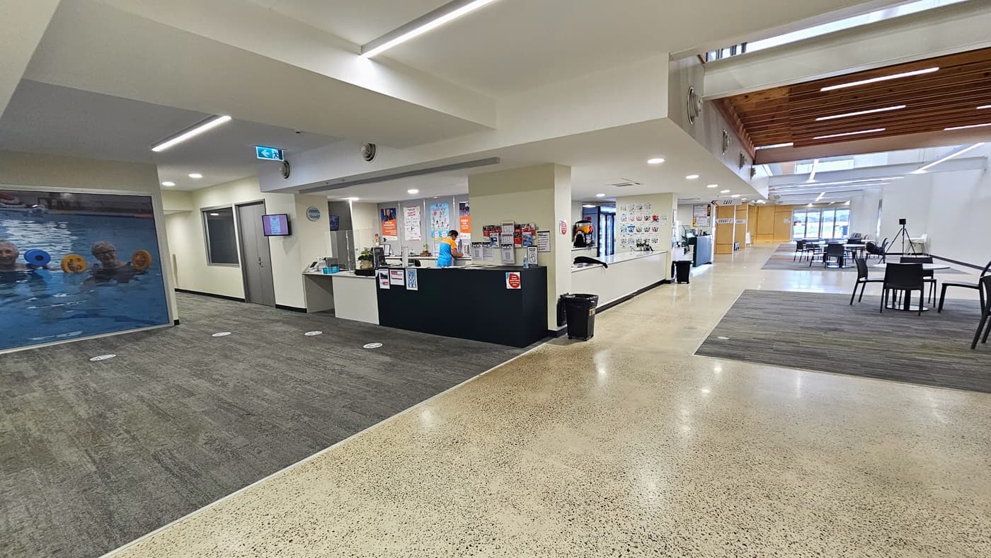 A closer look at the spacious corridor near the reception area. A sizable frame of two individuals in a pool is prominent on the left. Above a flat-screen TV on the left side of the reception area is an exit sign. The reception area features several posters on its desk and walls. An individual is noticeable inside the reception. Two black trash bins are situated on the right side. Further in the right area are black movable chairs and tables.