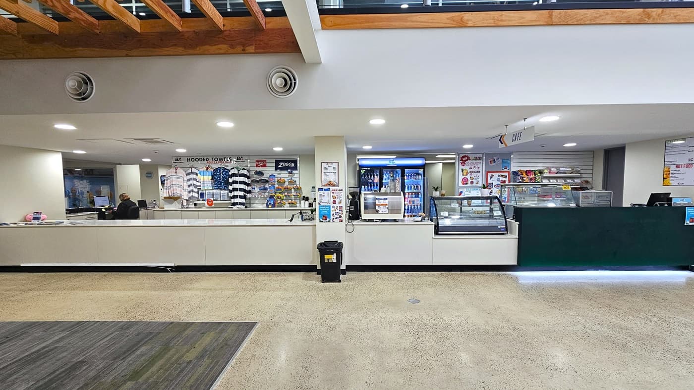 A closer look at the reception area. On the left side of the reception is a small retail store selling swimsuits, swimming goggles, etc. On the right is a glass display of varying cold beverages, snacks, and foods from the cafe. In the middle is a black trash bin. Two black speakers are affixed above the wall on the left.