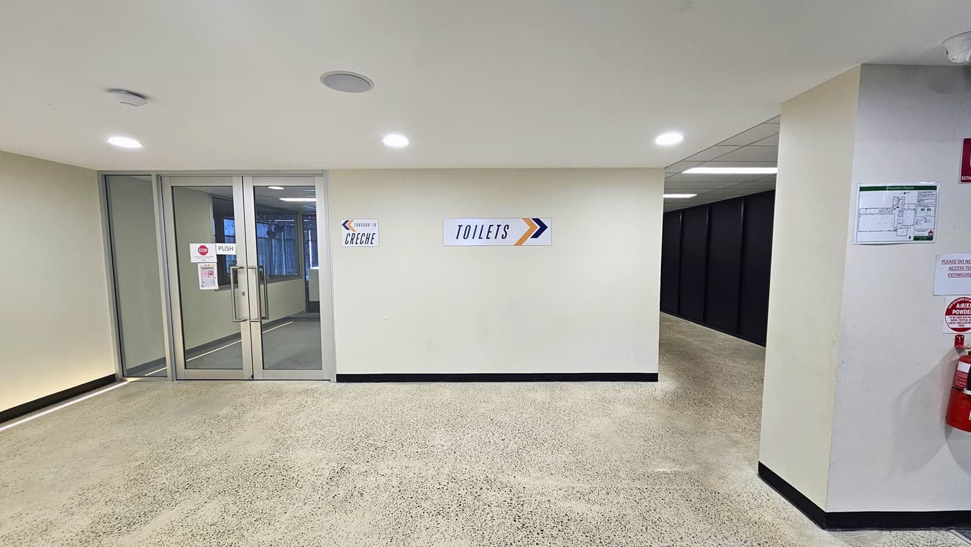 Pathway leading to the creche and to the toilet room. Signs that indicate specific directions of the creche and the toilet room are prominent on the wall in the middle. On its left, a manual glass door with small warning and directional signs on the glass is prominent. Beneath the CCTV on the right side is a layout poster of the facility, signs, and a fire extinguisher.