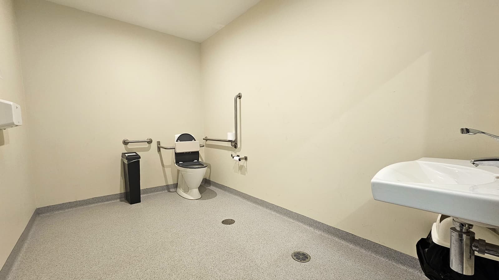 Inside the spacious accessible bathroom in Copper Coast Sport and Leisure Centre. The bathroom has a light brown natural stone flooring and a white wall. A thin black rubbish bin is placed on the left side of the toilet. Grab bars are on the back and both sides of the toilet. On the right is a hand wash basin. A hand sanitizer is affixed on the left wall.