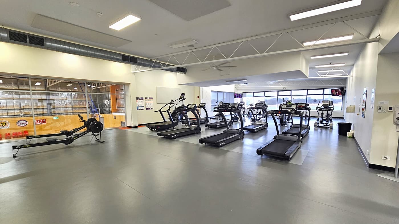 Half-open, double glass, manual outward-opening doors of the Cardio room. On the door is a small, black rubber mat. The room has glass windows with stainless steel frames. A ‘No Towel, No Train’ sign is placed above the PUSH sign on the glass door. Behind it is a red fire distinguisher mounted on the wall. Other small adverts can be found on the glass walls. Seen through the glass doors is a wooden cabinet on the left and the black stationary bikes on the right.