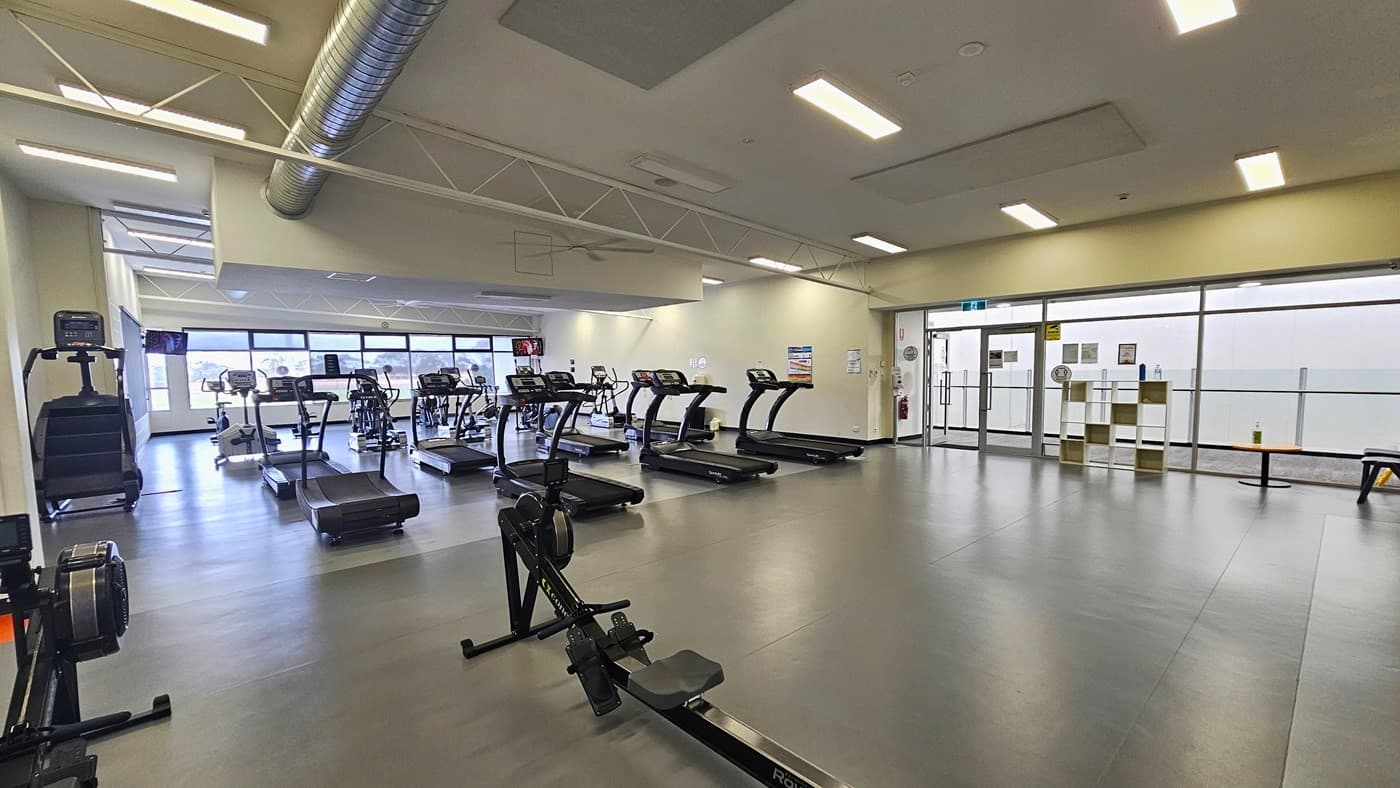 Back of the rows of black treadmills. Two black indoor rowing machines are available in the dark grey floor section. The upper part of the glass windows in front of the treadmills is tinted. Several small colorful adverts are affixed on the white wall and glass wall to the right. Near the manually operated glass doors are a wooden cabinet, an orange circular table, and black chairs. There is a large silver duct on the ceiling for the ventilation system.