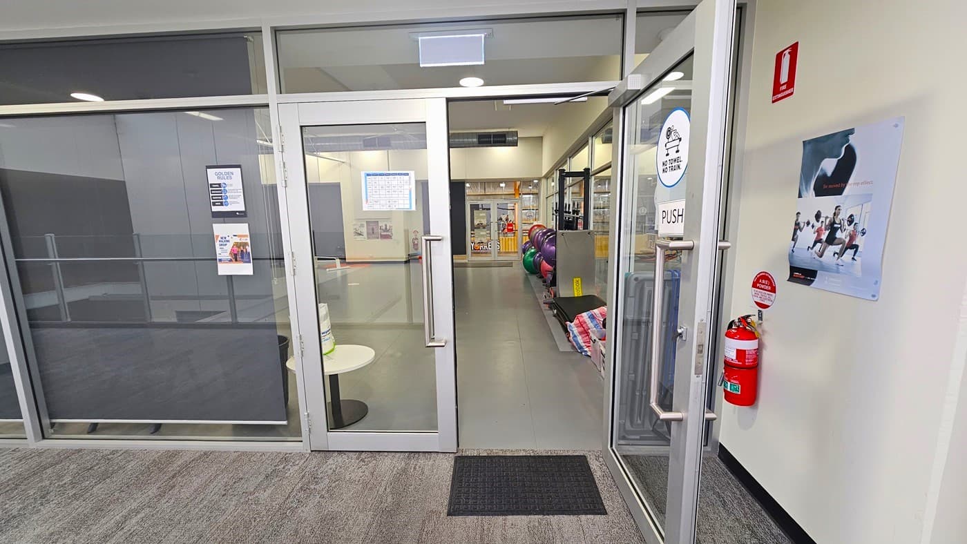 Half-open, double glass, manual outward-opening doors of the Group fitness room. On the door is a small, black rubber mat. The room has glass windows with stainless steel frames. A ‘No Towel, No Train’ sign is placed above the PUSH sign on the glass door. Behind it is a red fire distinguisher mounted on the wall. Other small adverts can be found on the glass walls. Seen through the glass doors are varying colors of exercise balls arranged against the right wall.