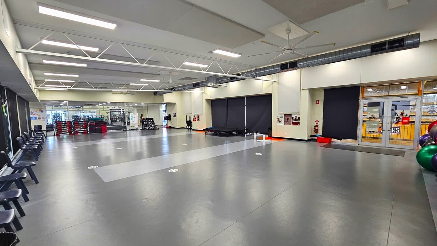 Inside the Group fitness room. The room has dark grey floors with two wide rectangular strips of light grey sections surrounded by white circles. Lined against the left wall and covered with black cloth are black monoblock chairs. To the back is a stack of black vertical steps with red step risers. On the black wall section to the right is a small black stage.