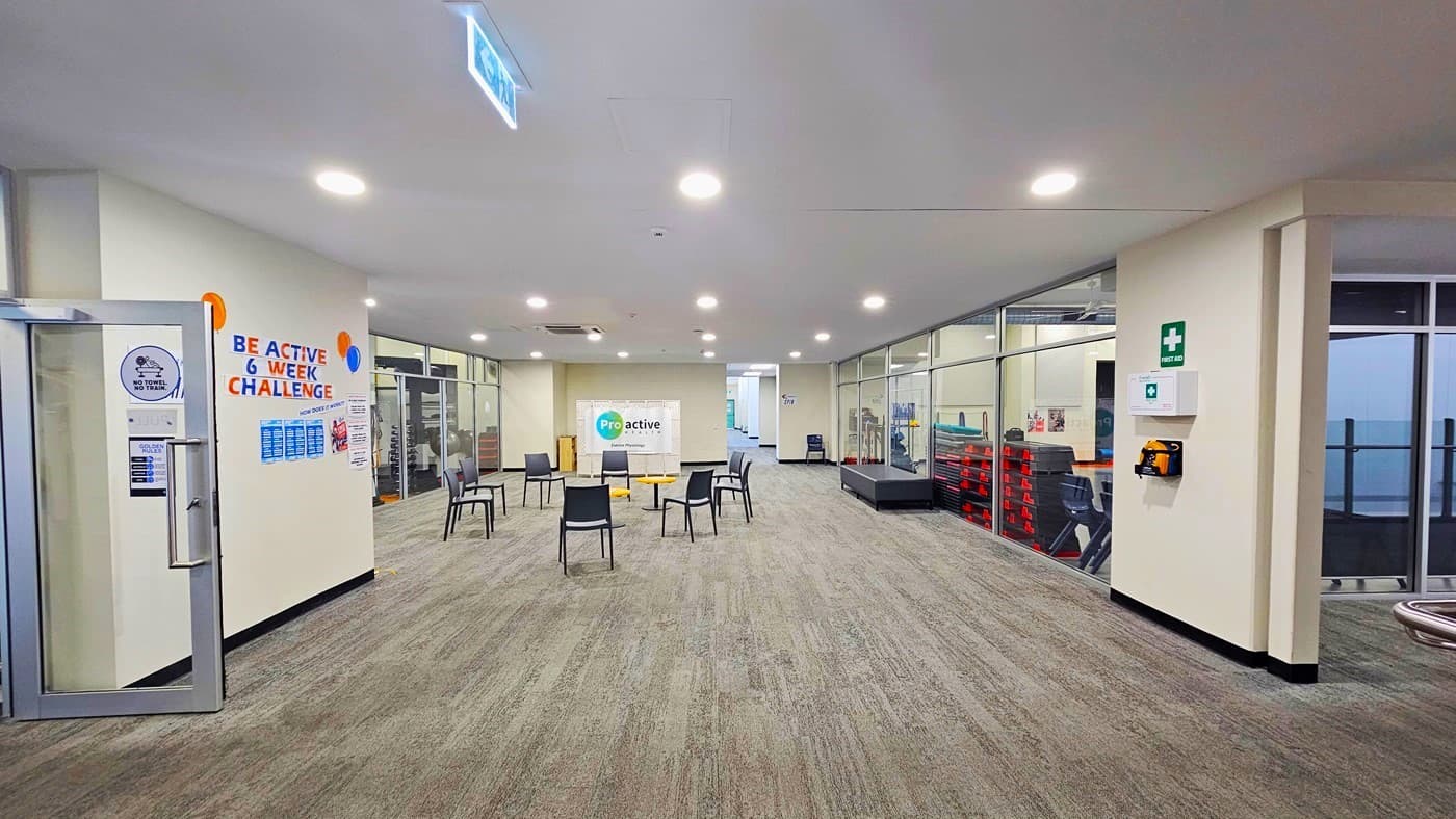 Rusty-looking floor area with seating in the middle. The seating area has black chairs around the yellow circular tables. On the left cream wall is an orange and blue ‘BE ACTIVE 6 WEEK CHALLENGE’ sign and balloons as well as small adverts. On the right wall is the white first aid kit box with a green ‘FIRST AID’ label and logo on top. Against the right glass wall is a soft black rectangular sofa. To the back is a white ProActive foldable floor signage.
