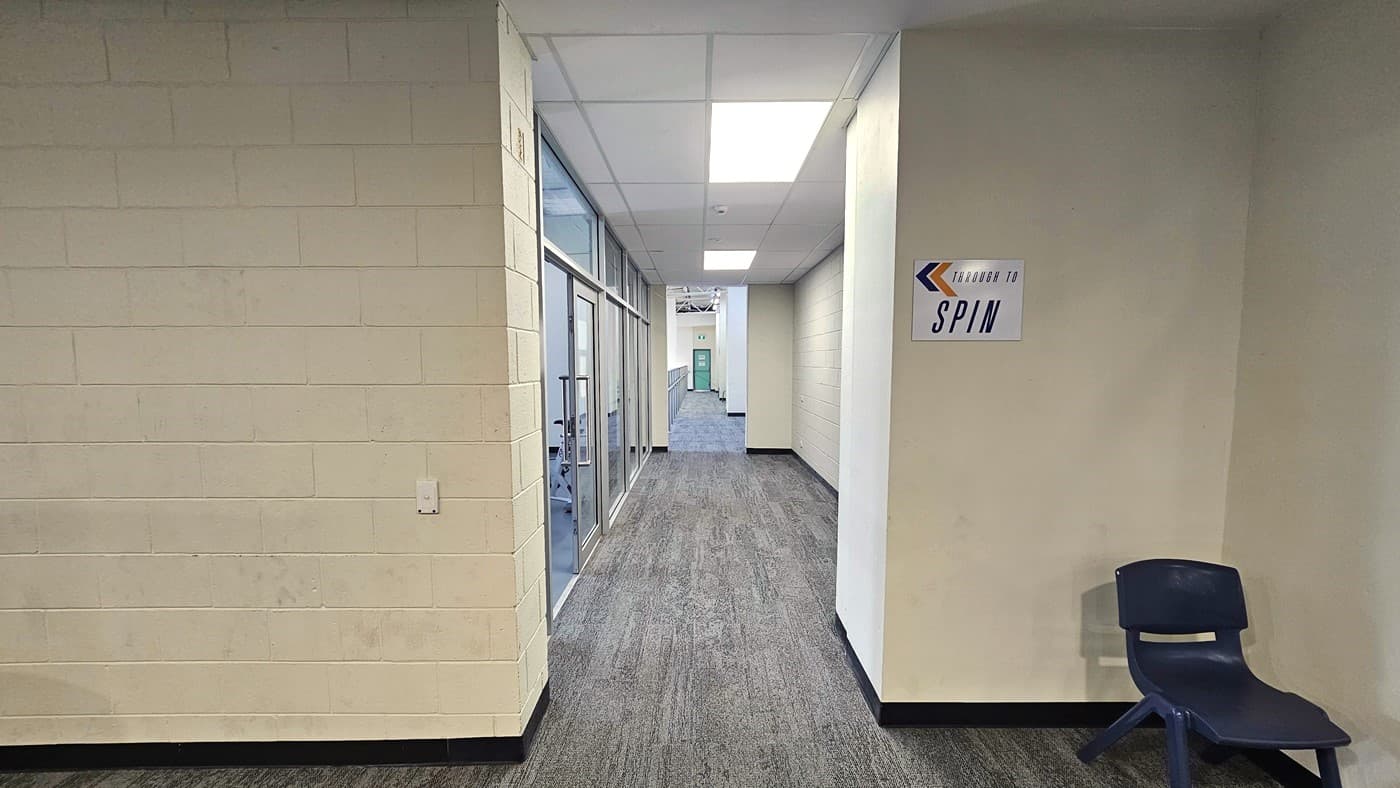 Narrow corridor leading to the Spin room. The corridor has a similar rusty-looking floor area, a white brick wall on the left, and a white smooth on the right. There is a white ‘Through to Spin’ sign with orange and blue arrows pointing to the left on the right white wall, and a black plastic chair is placed against the wall. The ceiling is white with tile-style fluorescent lighting. There is a green door at the end of the corridor.