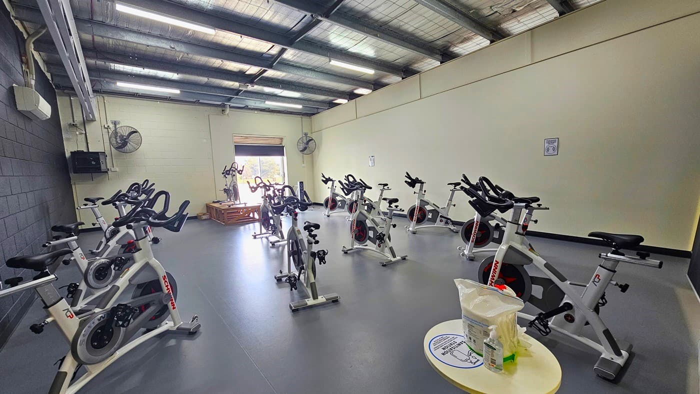 Inside the Spin room are white stationary bikes occupying the entire grey floor. The room has a left grey brick wall, a cream brick wall near the door, a smooth cream wall on the right, and a ceiling with black metal beams and silver insulators. There is a white split-type air conditioner on the grey brick wall. On the cream brick wall near the entrance door are two large wall-mounted black fans. On the left of the entrance is a single stationary bicycle placed above the wooden platform.