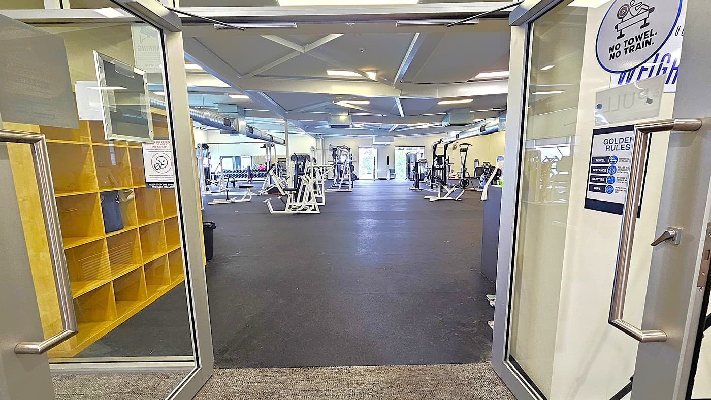 Entrance leading to the spacious Gymnasium. Signs are affixed to the glass of the manual sliding doors. Upon entering the facility, there is a yellow wooden cabinet positioned on the left side. The area is adorned with various exercise equipment and training machines.