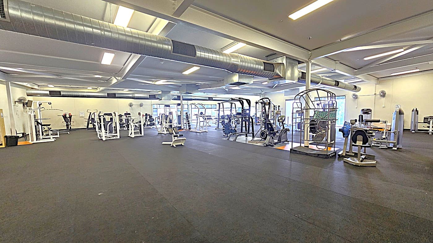 Another angle inside the Gymnasium. Two slim insulated beams are positioned overhead in the ceiling. Black and white workout machines and equipment are located on the right side and beyond. White walls in the gymnasium are adorned with mounted wall fans.