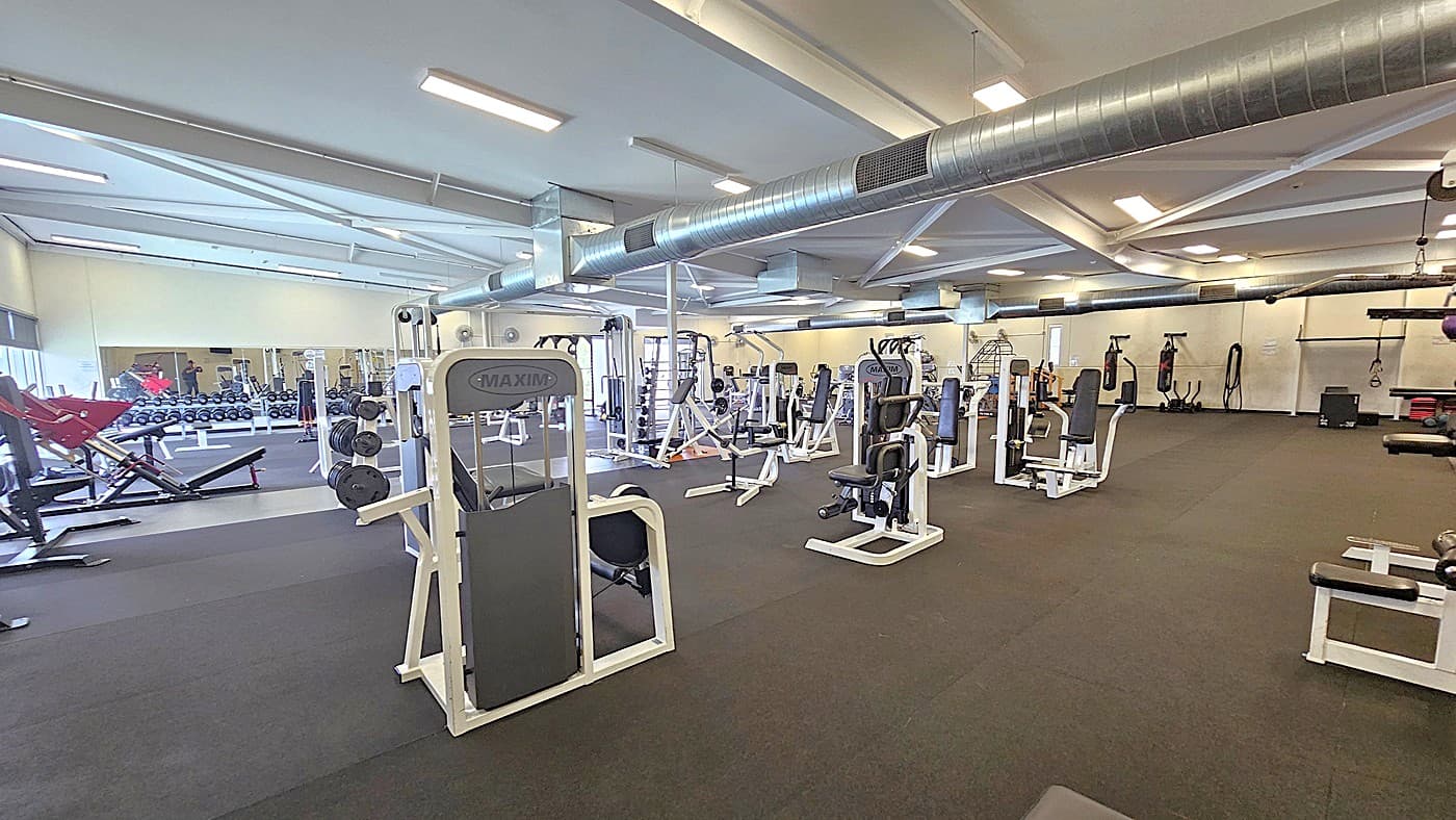 A closer look at the gym equipment. A broad mirror on the gym's walls reflects an array of dumbbells in different sizes on the left side. The gymnasium is filled with diverse black-and-white exercise machines and equipment. On the right side are two hanging black punching bags.