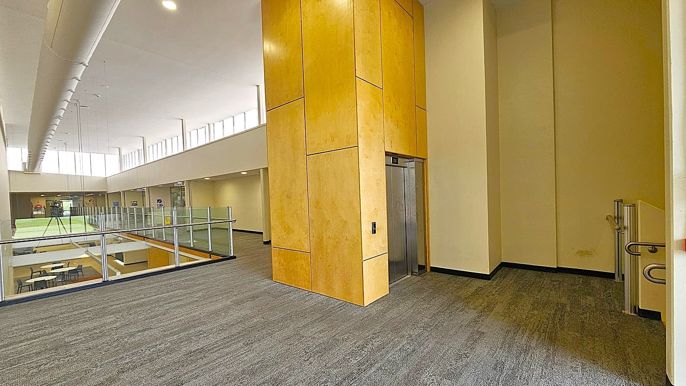 Walkway leading near a functional elevator. An elevator in the yellow-colored walls is situated right in front of the stairs on the right. The walkway’s floor surface features black and white vinyl flooring. White tables and black chairs are noticeable below on the left side.