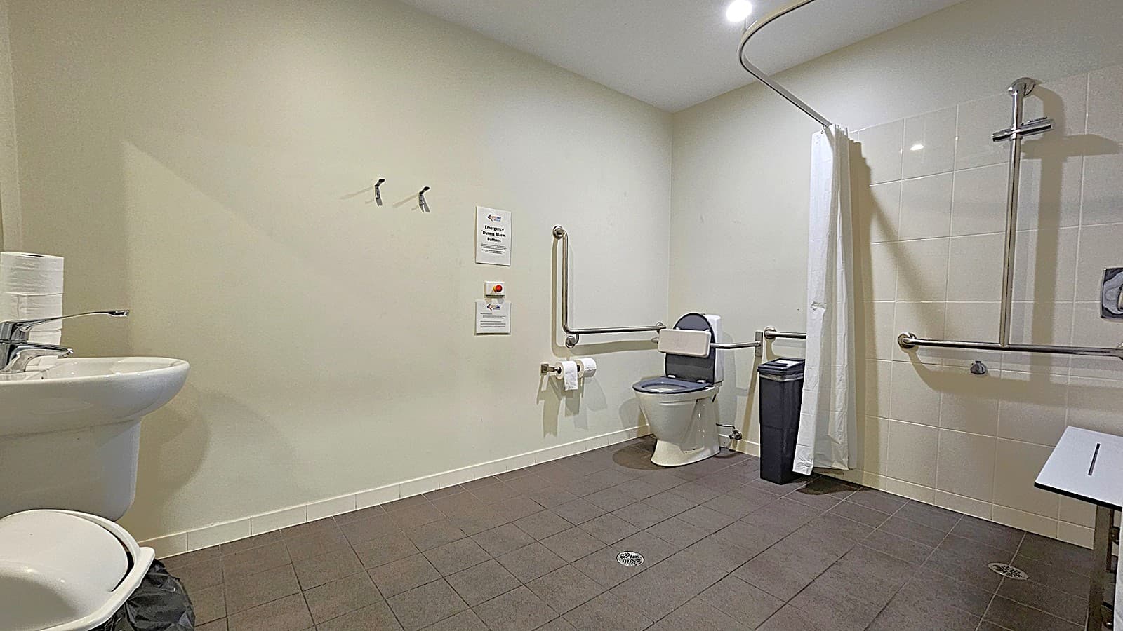 Inside the accessible toilet with a shower. The room features white walls and faded brown floor tiles. The shower area has a white curtain, a grab bar, and a small white bench on the right. On the left side of the toilet, two toilet roll holders with toilet paper are affixed to the wall. A black trash bin is placed on the right side of the toilet. Signs and a grab bar are above the toilet roll holders. A bathroom sink and another trash bin are on the left.