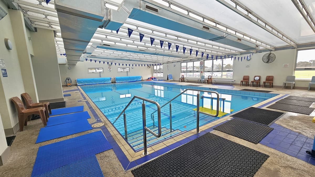 Entrance leading to the Hydrotherapy Pool. On the left side of the glass doors, a yellow warning sign that reads “slippery when wet” is affixed on the white walls. A blue floor mat is placed in the entryway. A brown chair sits on the left. Two floor mats are visible right after entering. Small posters are placed on both sides of the manual glass doors.