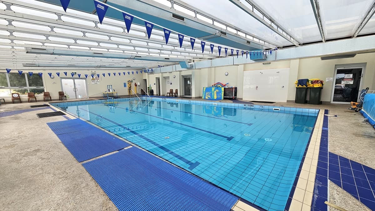 Another angle inside the pool. Blue pool flags are hanging above the ceiling. Black and blue anti-slip mats are situated on the left. Further ahead, on the left near the entrance, four brown chairs are placed. Two sizable black trash bins are on the right, next to another entrance. Next to the trash bins is a whiteboard.