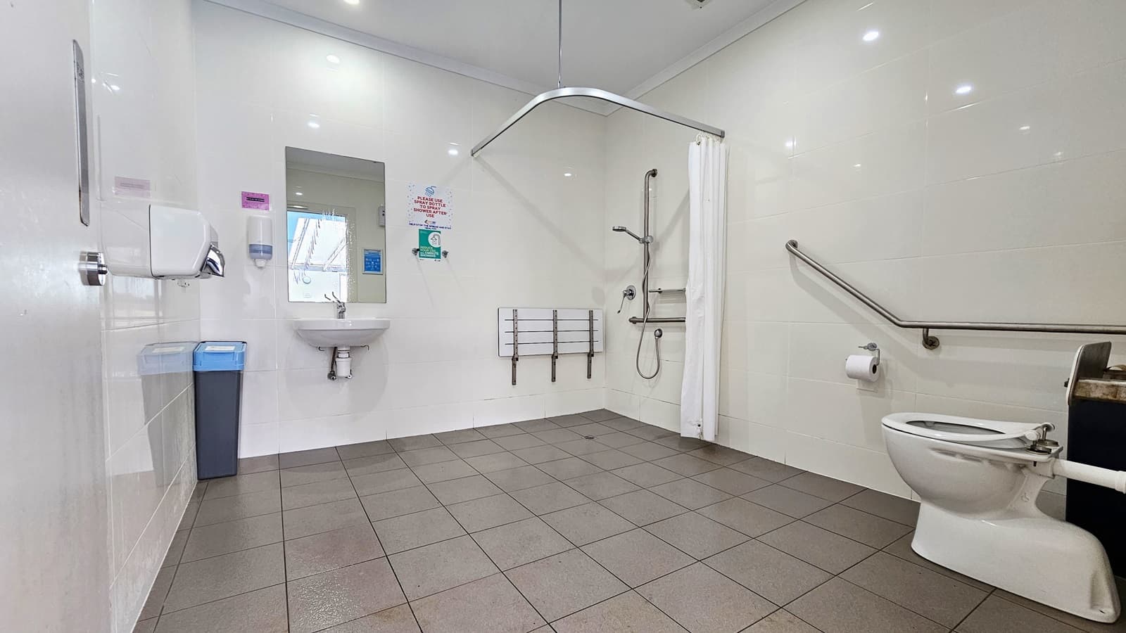 Inside the spacious accessible bathroom. The room features white walls and faded brown surface tiles. The toilet sits on the right, next to a black trash bin and a grab bar. Below the grab bar is where the toilet roll holder with toilet paper is situated. The shower area features a white shower curtain and a hand shower in a grab bar. On the left side of the grab bar is the small white bench. A bathroom sink and a mirror are affixed next to a trash bin and a hand sanitizer on the left.