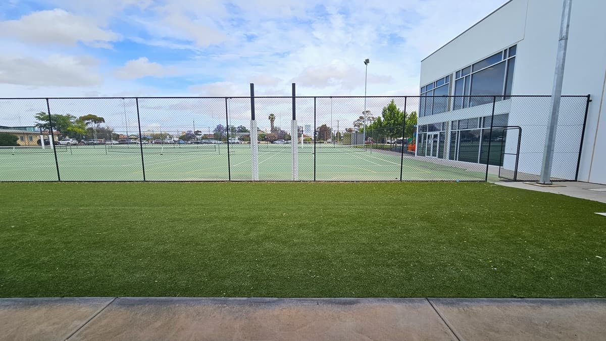 View of the outdoor tennis courts at Copper Coast Sport and Leisure Centre. The court is surrounded by a black metal chain-linked fence with an open door gate on the right side near a metal post. The court is green with white lines and has a black net in the middle. On the right side, there is a large white and grey industrial building with glass windows. Beyond is the car park filled with varying colors and brands of cars.