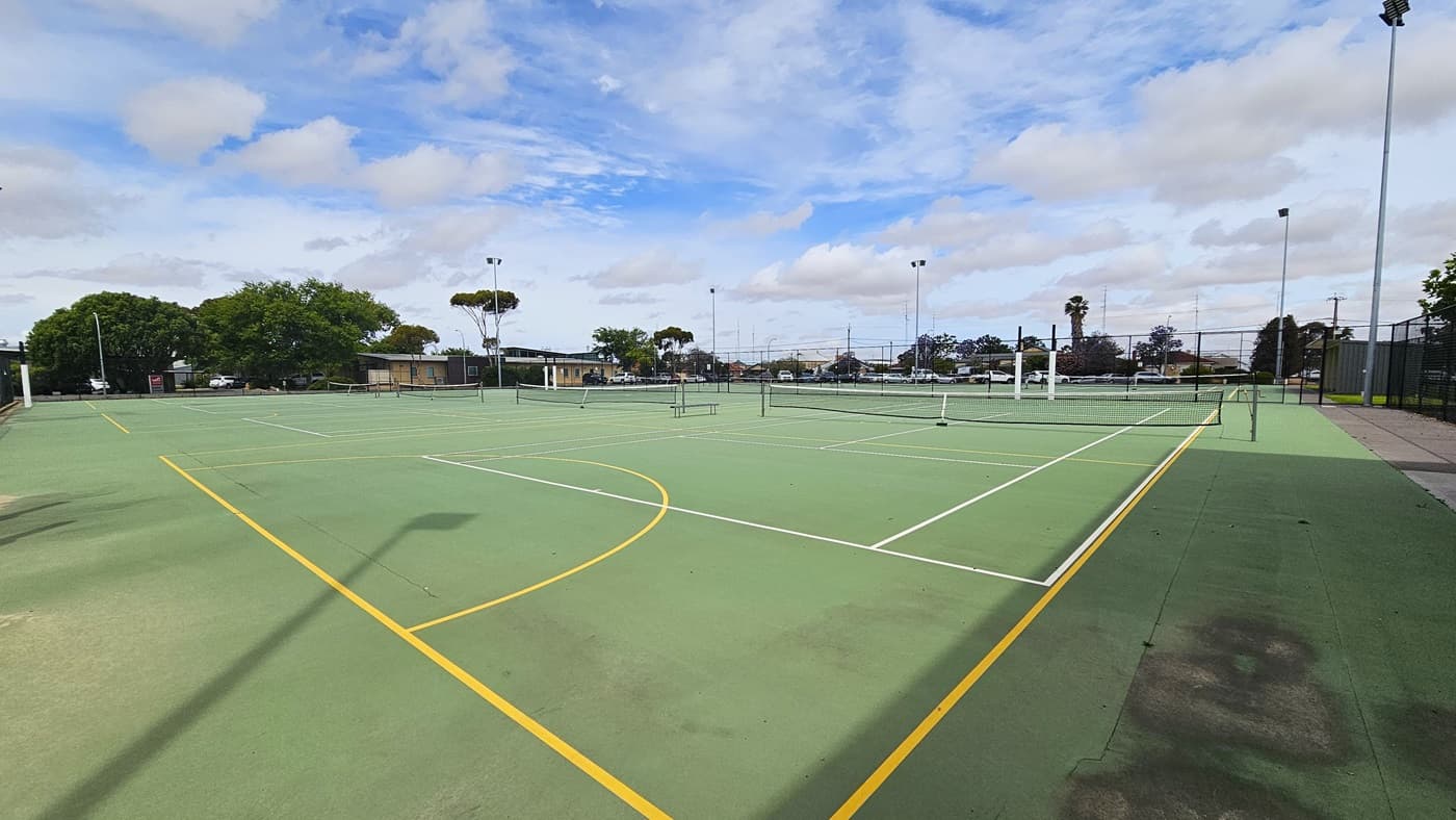 Outdoor tennis courts at Copper Coast Sport and Leisure Centre. The court is green with white and yellow lines and has a low black net in the middle. There is a cemented walkway on the right side connected to the next walkway to the back for the other set of tennis courts. Tall metal posts with lighting fixtures on top surround the entire perimeter of the court. Trees, residential buildings, and the car park can be seen from this position.