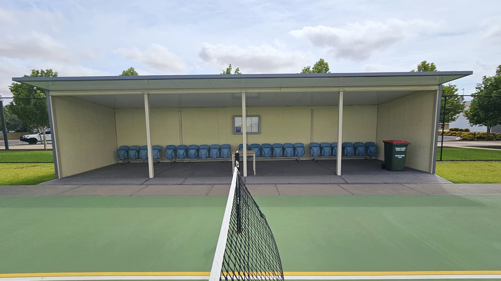 Sheltered seating area on the right side of the outdoor tennis court. The seating area is a rectangular structure with a flat roof and open front. The seating area has blue plastic chairs, a small table in the middle, a white scoreboard mounted on the back wall, and a green trash bin with a red lid on the right. The tennis court is green with white and yellow lines. The net is black and is stretched across the court. Behind the sheltered seating area are trees and a parking lot.