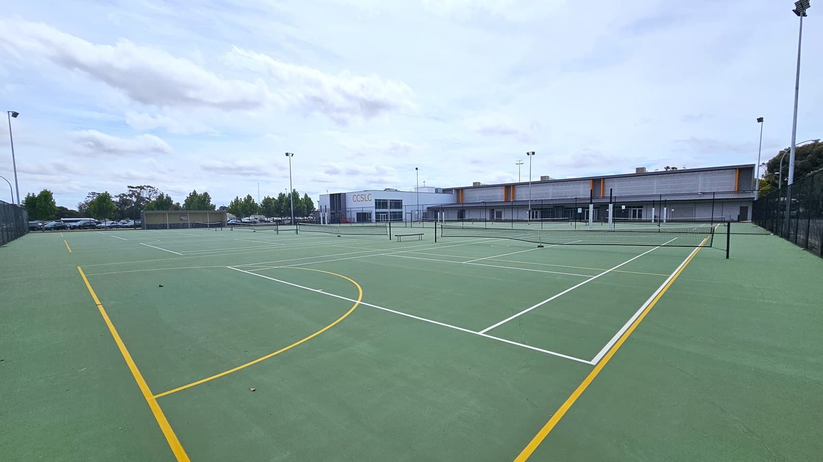 Another angle in one of the tennis courts. The court is green with white and yellow line ground markings. The court is surrounded by a black chain-linked fence and tall metal lamp posts. Further to the left is the parking lot with a number of cars parked. To the back is a large, white, and grey industrial building with a bold yellow-orange “CCSLC” sign on the front of the façade. Beside is a grey-tiled building with orange wall tubular accents.
