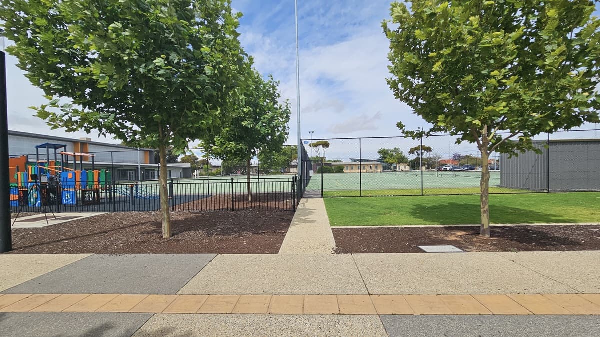 External entry to the tennis court for after-hours access directly from the car park. A concrete pathway connected to the brick path from the building entrance leads towards the gate of the black chain-linked fence enclosing the entire perimeter of the tennis courts. On the left is a playground with colorful play features with a low set metal bar fence, while on the right is the sheltered seating with grey corrugated wall. Trees with abundant foliage provide shade to the pathway.