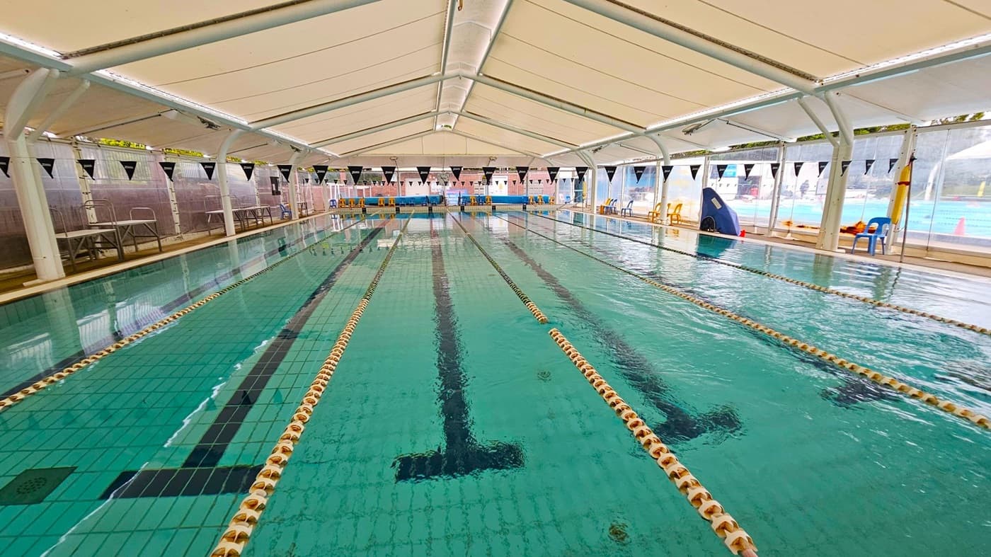 An indoor swimming pool with multiple lanes, separated by floating lane dividers. The pool is covered by a white tent-like structure with a high ceiling. On the left and right sides, there are transparent walls with black triangular flags hanging above. Several plastic chairs in various colours are placed along the sides. The floor of the pool is tiled in green with black lane markings.