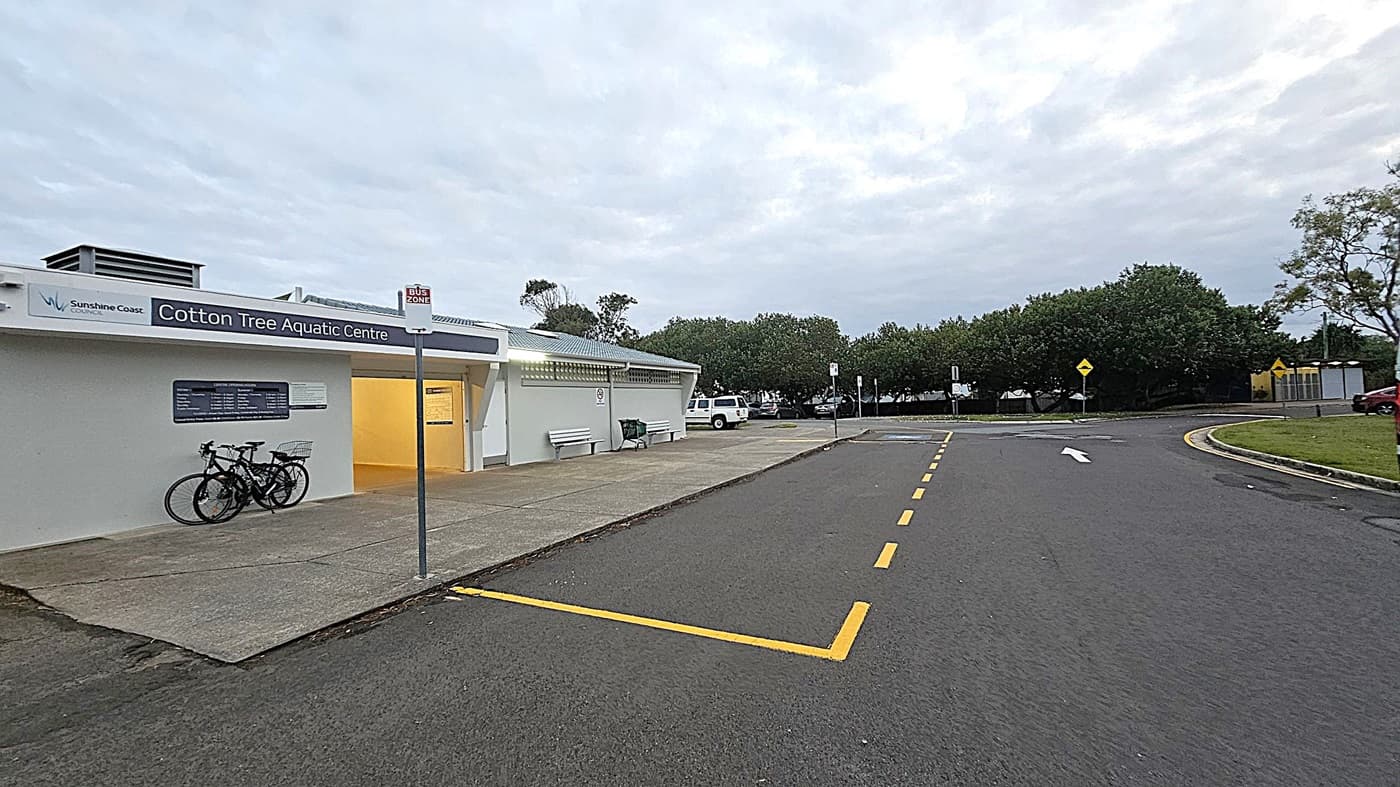 A car park with two vehicles in designated disabled parking spaces. On the left, a black Mazda with a visible licence plate. To the right, a grey SUV. Behind the cars, a low building with a sign reading "Clifton Hill Aquatic Centre" is partially visible. Surrounding the area are green bushes and trees. A streetlamp is lit near the left side, and the ground is covered with dark asphalt.