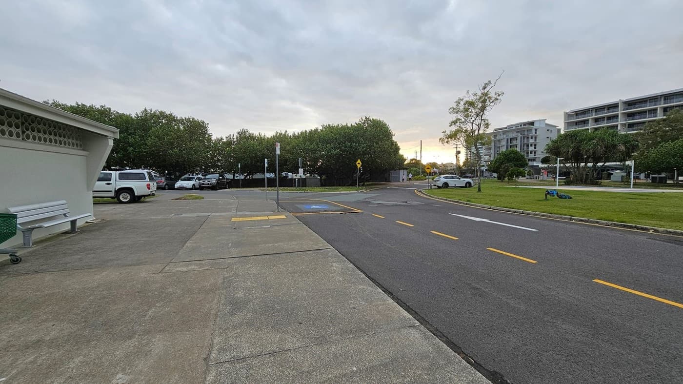 A paved area with a bench and a green bin on the left, next to a building with a perforated wall. Several parked cars are visible in the background, partially obscured by trees. On the right, a road with yellow dashed lines runs alongside a grassy area with a few trees and a white car. In the distance, there are multi-storey buildings. The sky is overcast, and the scene appears to be at dusk or dawn.