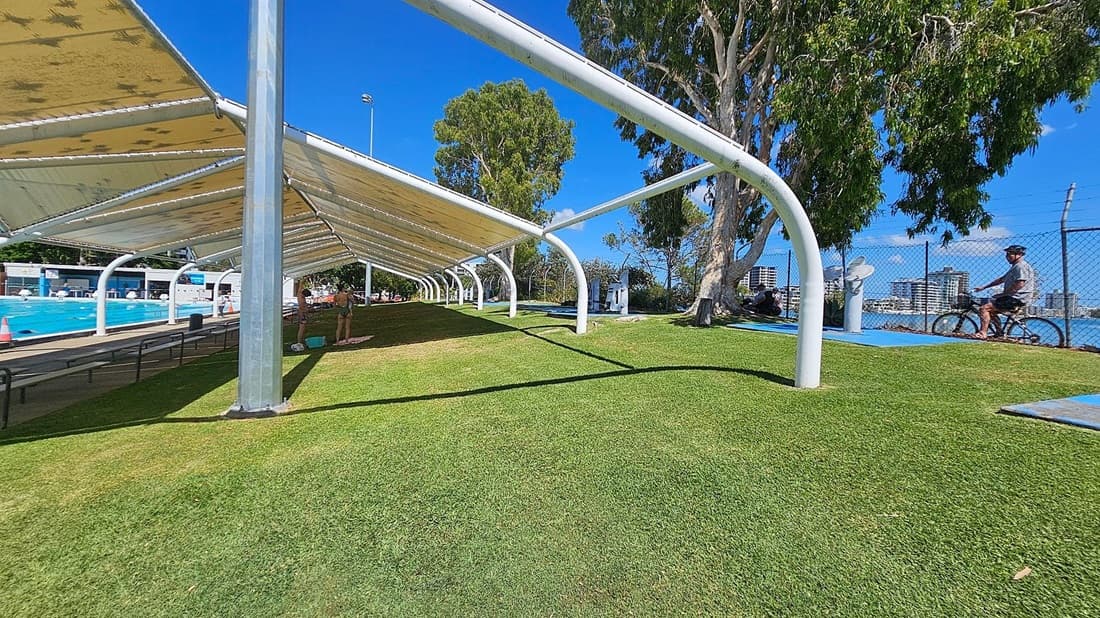A grassy area with a large, white metal canopy structure on the left, providing shade. Underneath, there are benches and a few people standing near a swimming pool. On the right, a person is cycling along a path next to a chain-link fence. Tall trees are scattered throughout, and buildings are visible in the background. The sky is clear and blue.