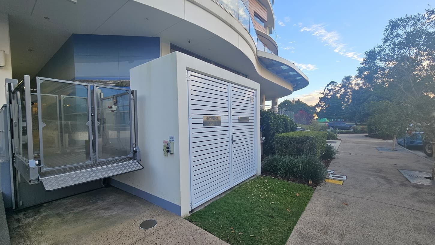 On the left, there is a metal platform lift next to a building with blue-tinted glass windows. In the middle, there is a white structure with horizontal slats and a small access door. On the right, there is a pathway bordered by green shrubs and trees, leading to a parking area with a few cars. The floor is a mix of concrete and grass, and the ceiling is part of the building's overhang.