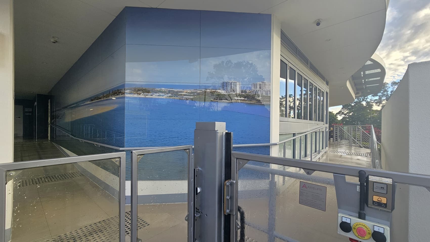 On the left, there is a corridor with a tiled floor and a wall featuring a large mural of a coastal scene. In the middle, the mural continues, showing a blue ocean and cityscape. On the right, there is a glass railing and a control panel with buttons and a sign. A stair with handrails is positioned on the far right. The floor is concrete with truncated domes, and the ceiling is smooth and white.