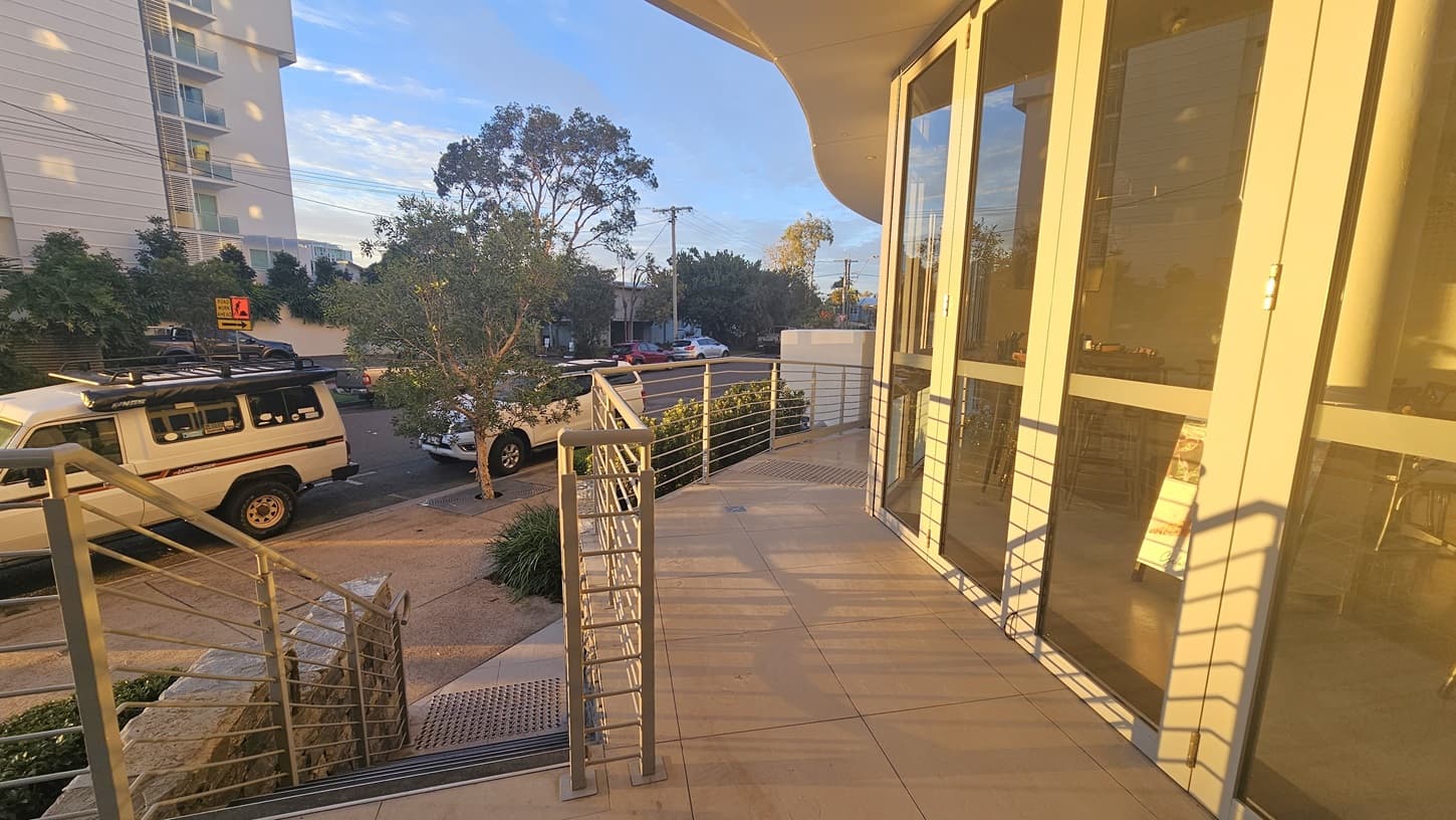 On the left, there is a metal railing alongside a ramp and steps leading down to a pavement. Truncated domes are installed on the floor before the steps. In the background, there are multi-storey buildings, parked cars, trees, and a building with white walls. On the right, there are large glass doors reflecting the surroundings, part of a building with a curved overhang. The floor is tiled, and the ceiling is smooth and white.