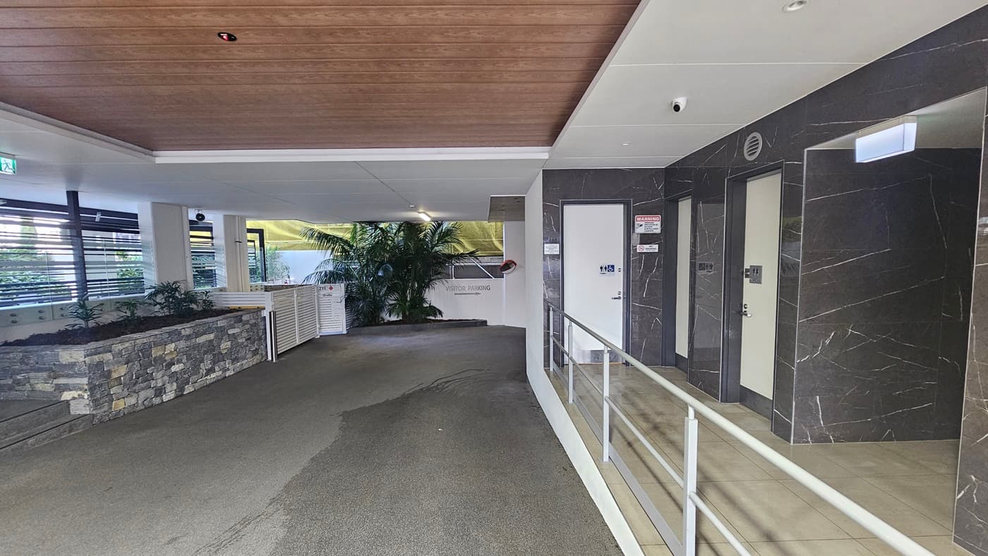 On the left, there is a stone planter with greenery and a white railing. In the middle, there is a pathway leading to a white wall with a large plant in front of it. On the right, there are two lift doors with control panels, set in a dark marble wall. The floor is a mix of concrete and tiled surfaces, and the ceiling is divided into sections with white panels and wooden panels.