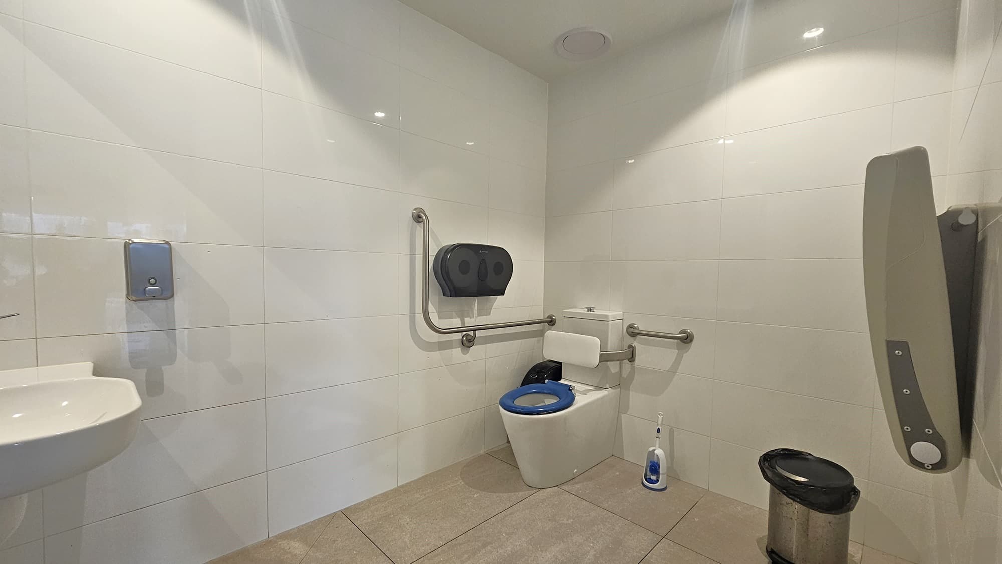 On the left, there is a white sink with a soap dispenser mounted on the tiled wall. In the middle, there is a toilet with a blue seat, flanked by metal grab bars and a toilet paper dispenser above it. On the right, there is a wall-mounted baby changing station and a small bin on the floor. The floor is tiled in a light grey colour, and the ceiling is white with recessed lighting.
