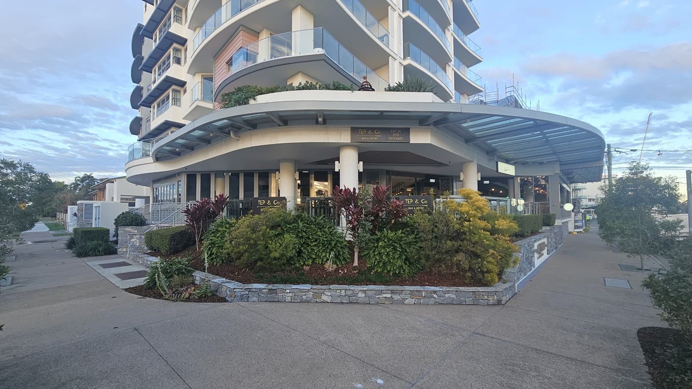 A multi-storey building with curved balconies on the left, featuring a ground-floor café with outdoor seating. The right side has a pathway leading to the entrance, bordered by landscaped greenery. In the middle, there are large windows and a glass canopy extending from the building. The foreground has a concrete pavement with a stone planter filled with various plants and shrubs.