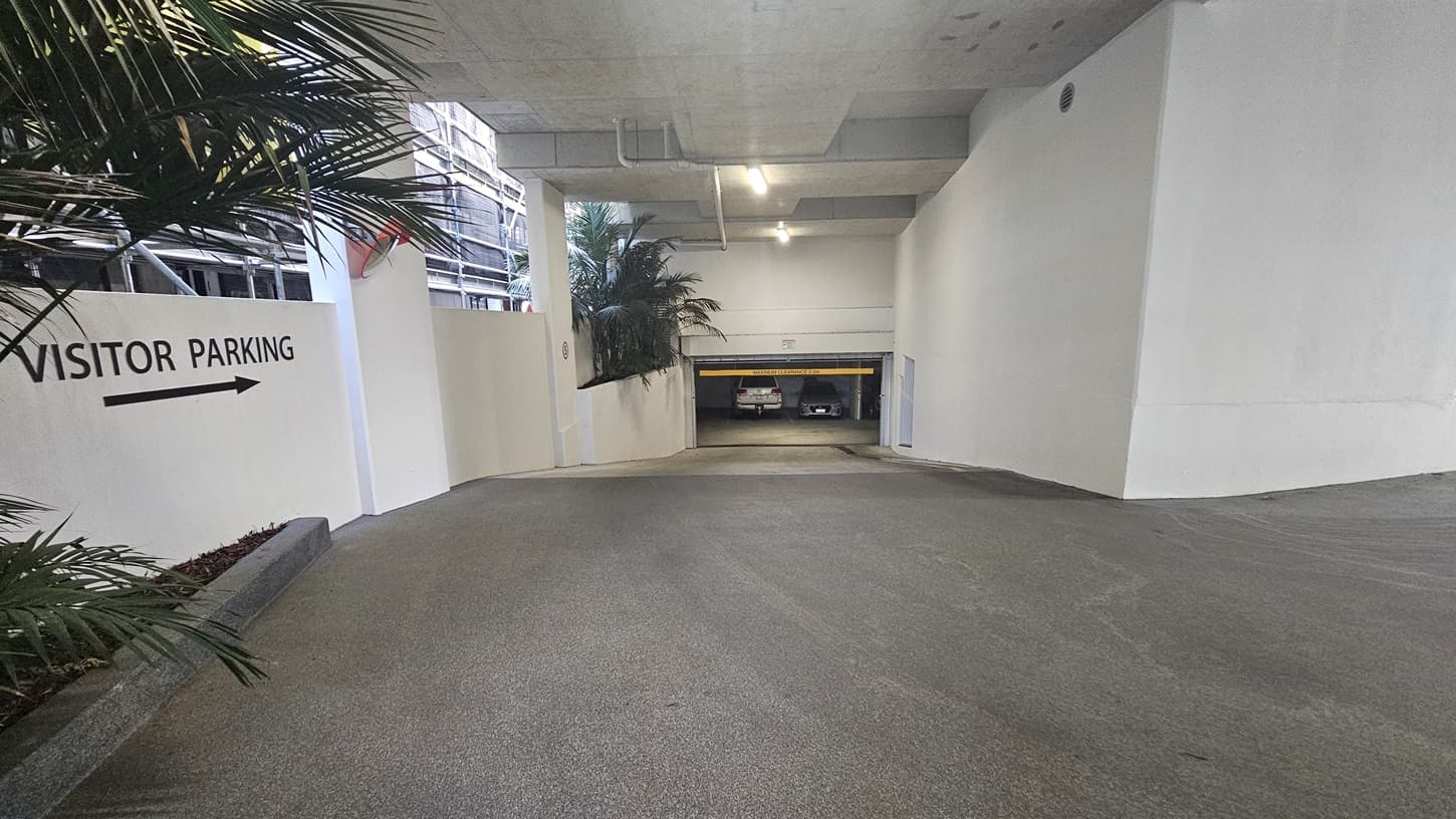 On the left, there is a white wall with a "Visitor Parking" sign and an arrow pointing right, next to a planter with tall palm plants. The right side has a plain white wall. In the middle, there is a sloped driveway leading down to an underground parking area with a yellow height restriction bar. The background includes parked cars and more palm plants. The floor is smooth concrete, and the ceiling is also concrete with exposed pipes and recessed lighting.