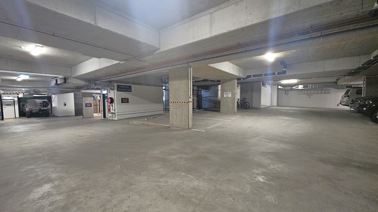 On the left, there is a parked white vehicle near a wall with a sign and a door leading to an exit. The right side has several parked cars and bicycles against the wall. In the middle, there is a concrete pillar with a height restriction bar and a sign indicating parking directions. The background includes more parking spaces and a wall with pipes. The floor is smooth concrete, and the ceiling is also concrete with exposed pipes and fluorescent lighting.