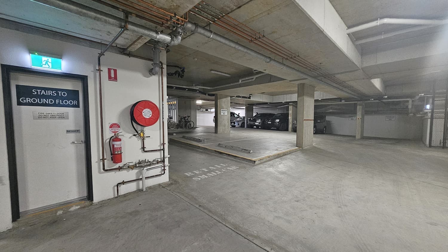 On the left, there is a door labelled "Stairs to Ground Floor" next to a fire hose reel and an extinguisher mounted on the wall. The right side has several parked cars and bicycles near concrete pillars. In the middle, there is a ramp leading to another level of the car park. The background includes more parking spaces and a wall with a sign indicating "Keep Area Clear." The floor is smooth concrete, and the ceiling is concrete with exposed pipes and fluorescent lighting.