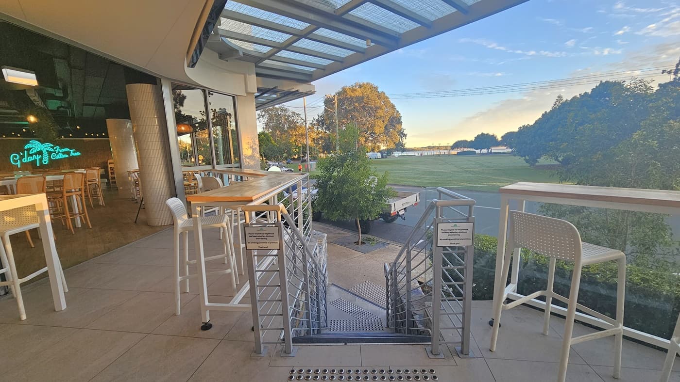 On the left, there is an outdoor seating area with high tables and stools, adjacent to a building with large windows and a neon sign inside. The right side has more high tables and stools, overlooking a grassy area with trees. In the middle, there is a concrete staircase with handrails leading down to a lower level. Truncated domes are installed before and after the stairs. The background includes a road, more trees, and a distant view of water.