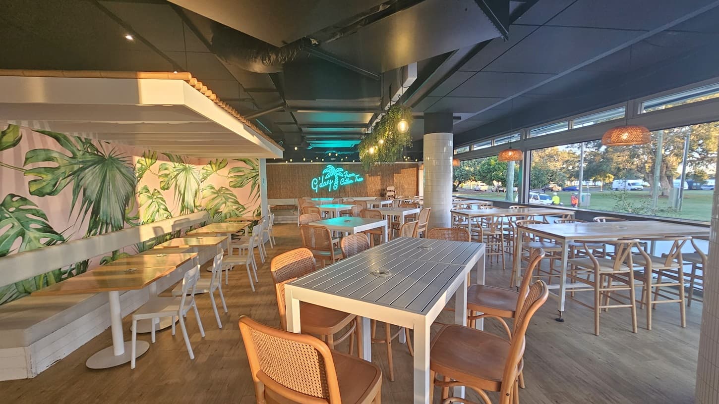 On the left, there are open glass doors leading into a café with white tiled walls and shelves holding various bottles and jars. In the middle, a counter with a cash register and a display of pastries is visible, with a hanging plant above it. On the right, more open glass doors reveal additional seating with white chairs and tables. The floor is wooden, and the ceiling has a mix of white tiles and dark panels with recessed lighting.