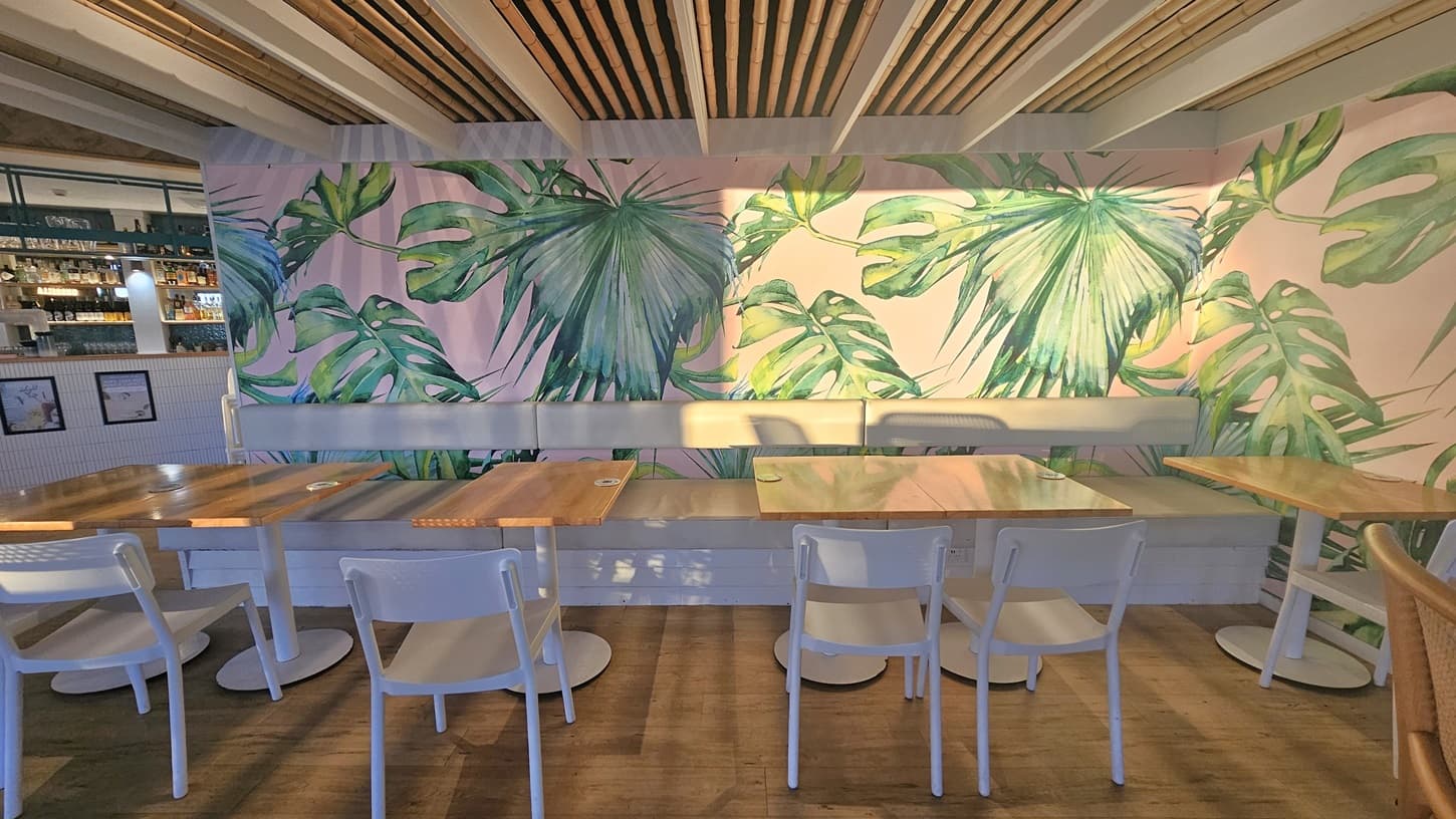 On the left, there are open glass doors leading to an outdoor area with trees and a grassy field visible. In the middle, a high wooden table with stools is positioned against a wall with a neon sign reading "Just Here For The Umbrella Drinks." On the right, there is a counter with a display of pastries, a coffee machine, and hanging plants above. The floor is wooden, and the ceiling has dark panels with recessed lighting and exposed ductwork.